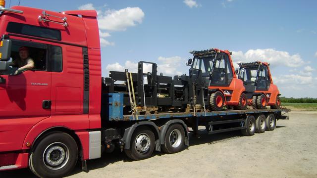 Czerwony ciągnik siodłowy MAN TGA 480 przewozi na platformie dwa pomarańczowe wózki widłowe Linde oraz czarne metalowe konstrukcje. Widoczny kierowca w kabinie.