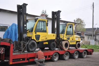 Dwa wózki widłowe Hyster, zabezpieczone łańcuchami, przewożone na niskopodwoziowej platformie transportowej. Jeden z wózków ma żółte pasy ostrzegawcze na bokach.
