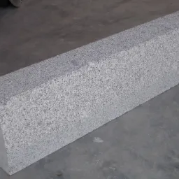 Szara kostka granitowa o prostokątnym przekroju, leżąca na szarym betonowym podłożu, z fragmentem opony w tle, prezentująca fakturę kamienia.