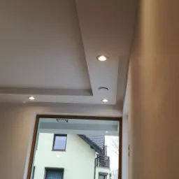 Nowoczesny sufit podwieszany z oświetleniem punktowym w jasnym wnętrzu, widok na dom przez okno.