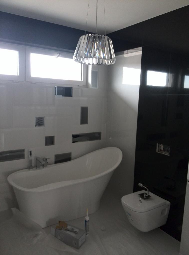 Nowoczesna łazienka z wolnostojącą wanną, podwieszaną toaletą i czarno-białymi płytkami, oświetlona kryształowym żyrandolem. Na podłodze widoczne narzędzia i opakowania po materiałach budowlanych.