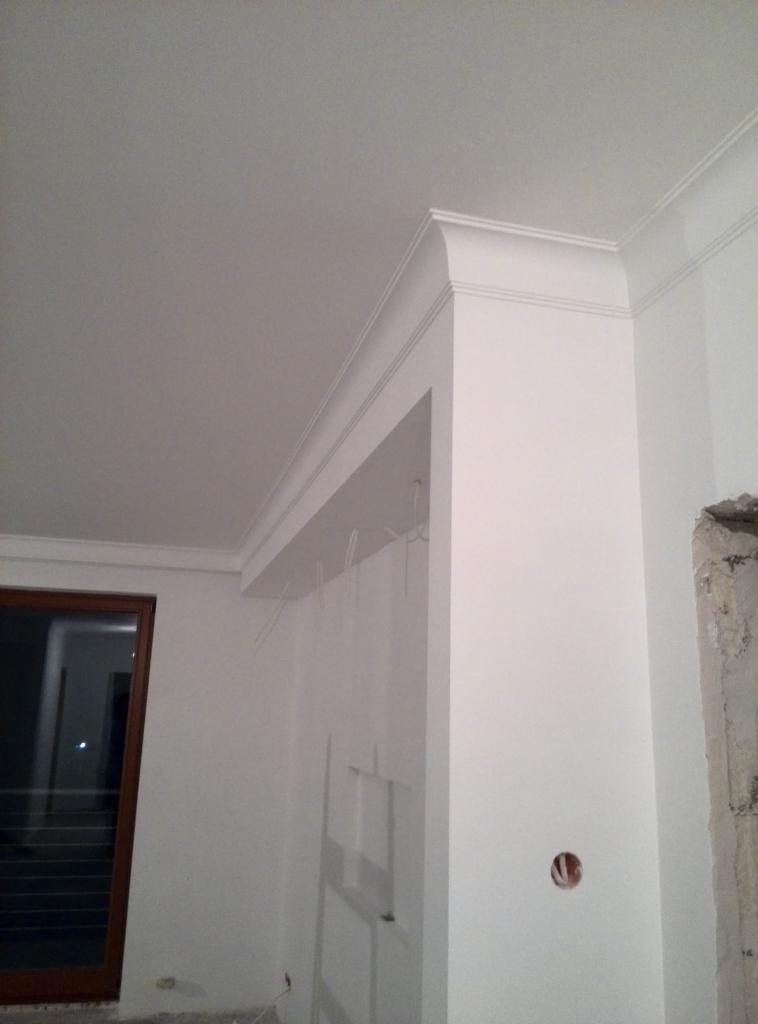 Wnętrze pokoju w trakcie prac wykończeniowych, widoczne białe ściany z ozdobnymi listwami przysufitowymi, surowy fragment ściany obok drewnianych drzwi i wystające przewody elektryczne.