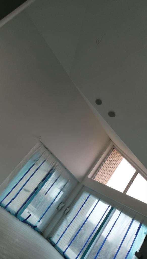 Białe wnętrze w trakcie zabezpieczania folią malarską okien i podłogi przed malowaniem, widoczne dwa okrągłe punkty oświetleniowe w skośnym suficie.