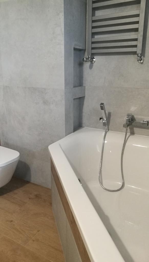 Wykończona łazienka w szarościach z drewnopodobną podłogą, widoczna narożna wanna z chromowaną armaturą, grzejnik drabinkowy i toaleta w tle.