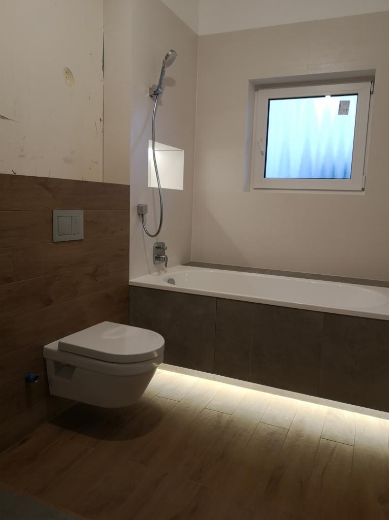 Nowoczesna łazienka w trakcie wykańczania: podwieszana toaleta, wanna obudowana szarymi płytami, podświetlana listwa przypodłogowa, drewnopodobne płytki na podłodze i częściowo na ścianie, widoczne...