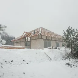 Surowa bryła domu w trakcie budowy, z widoczną drewnianą konstrukcją dachu bez pokrycia, otoczona śnieżnym krajobrazem i ośnieżonymi drzewami.