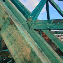 RADOSŁAW PEPLIŃSKI Cieśla Kaszuby - Fragment konstrukcji dachu z impregnowanego drewna w odcieniach zieleni, widok z dołu na tle błękitnego nieba z chmurami, detale połączeń ciesielskich.