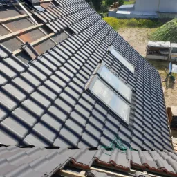 Montaż ciemnoszarej dachówki ceramicznej na łatach z widocznymi oknami dachowymi Velux i kominem, częściowo zakończony, z widokiem na okoliczne budynki i zieleń w tle.