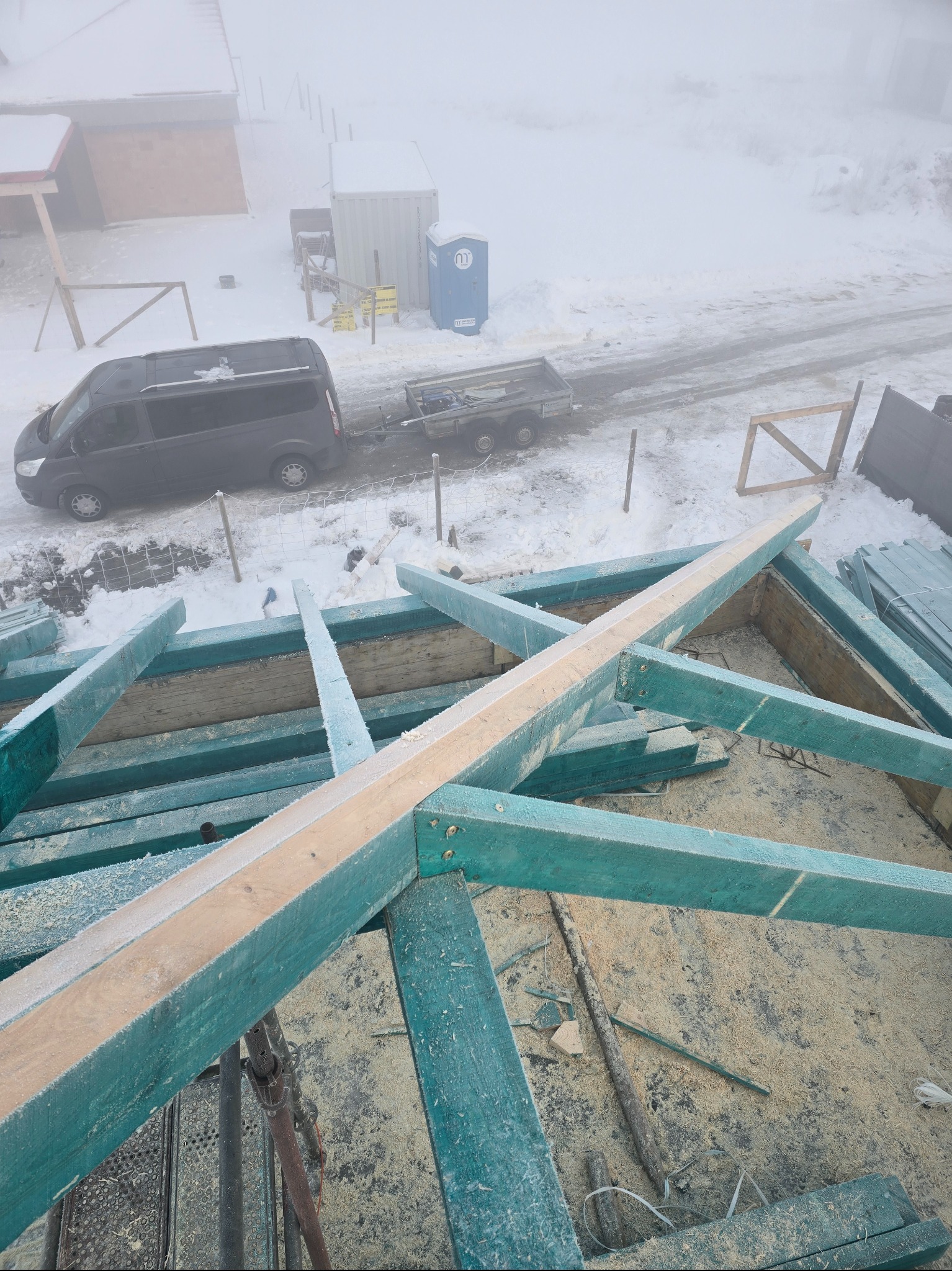 Wznoszenie więźby dachowej z impregnowanego drewna w zimowej scenerii. Widoczny fragment konstrukcji, samochód dostawczy i przenośna toaleta w tle, wszystko spowite mgłą.