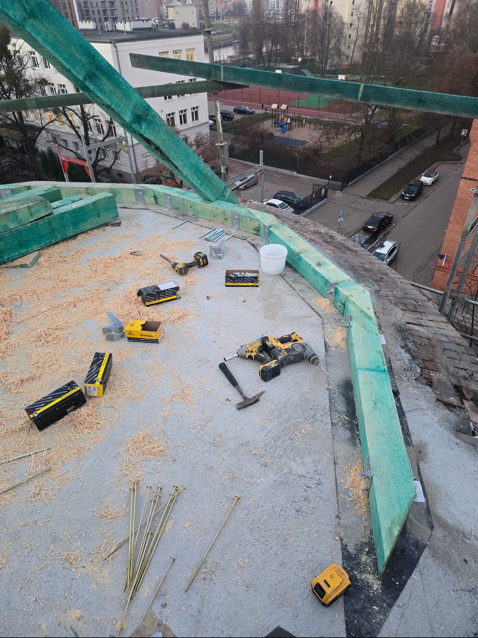 Montaż okrągłej konstrukcji dachu z impregnowanego drewna, wiertarki, gwoździe i wióry na płaskiej powierzchni dachu z widokiem na miasto w tle.