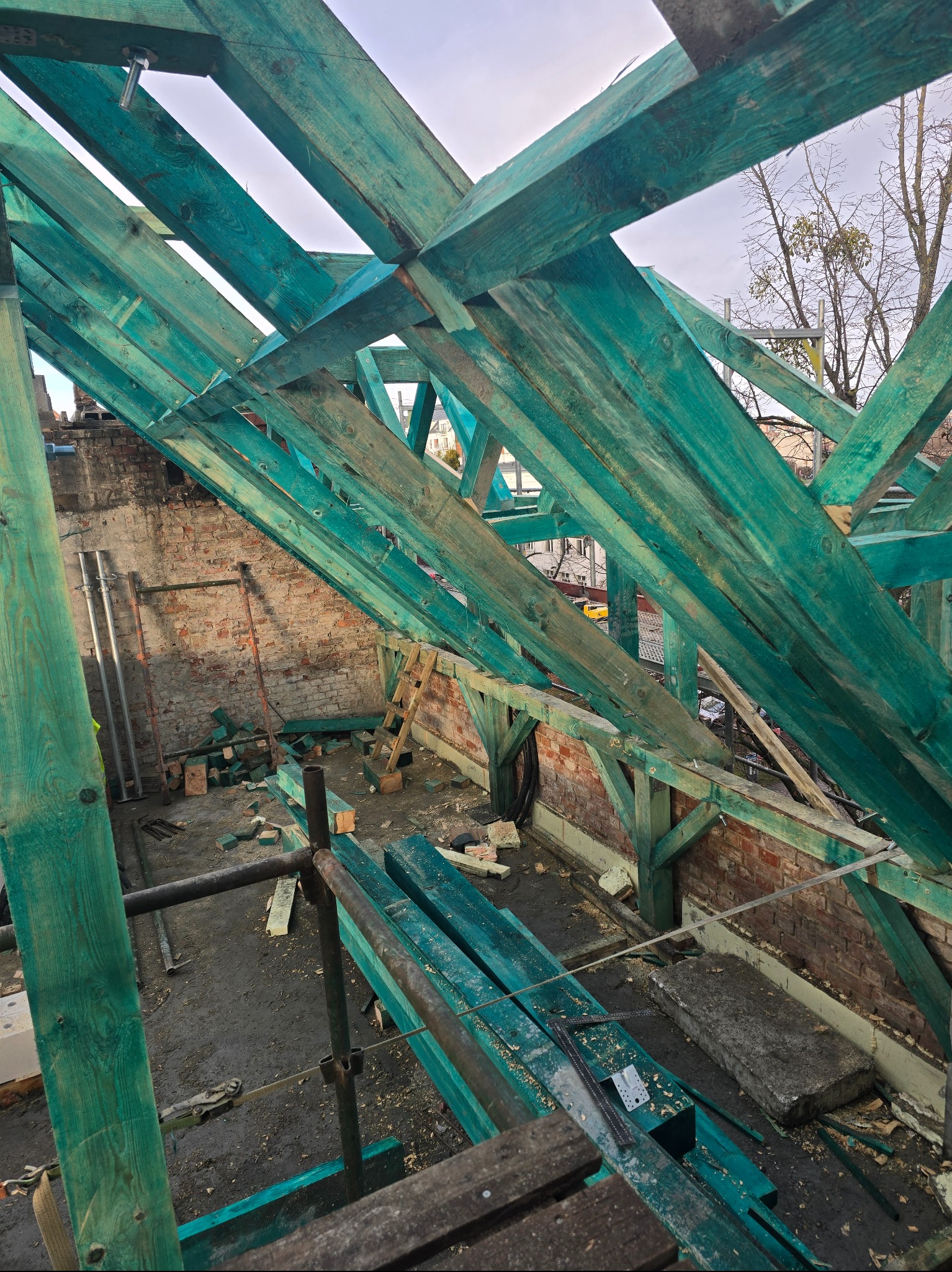 Turkusowa, drewniana konstrukcja dachu w trakcie budowy, widok z góry. Fragment muru z cegły w tle. Elementy rusztowania widoczne na pierwszym planie.