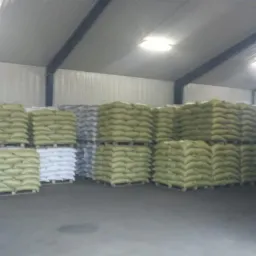 Magazyn wypełniony paletami z workami. Widoczne rzędy ułożonych worków w kolorach beżowym i białym, pod metalowym dachem magazynu z oświetleniem.