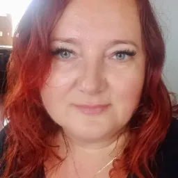 Portret kobiety z rudymi, kręconymi włosami, błękitnymi oczami i sztucznymi rzęsami, ubrana w czarną bluzkę, z delikatnym złotym łańcuszkiem z literką na szyi, na rozmytym tle wnętrza.