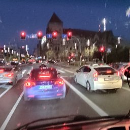 TW - Nocny widok z wnętrza pojazdu na ulicę miejską z samochodami stojącymi na czerwonym świetle, widoczne budynki w tle i krople deszczu na szybie.