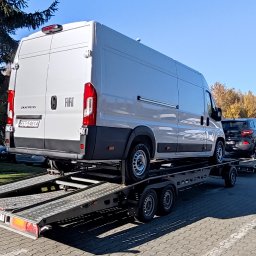 TW - Biały Fiat Ducato na platformie lawety z czarnym samochodem osobowym za nim, w tle drzewa i inne pojazdy na parkingu