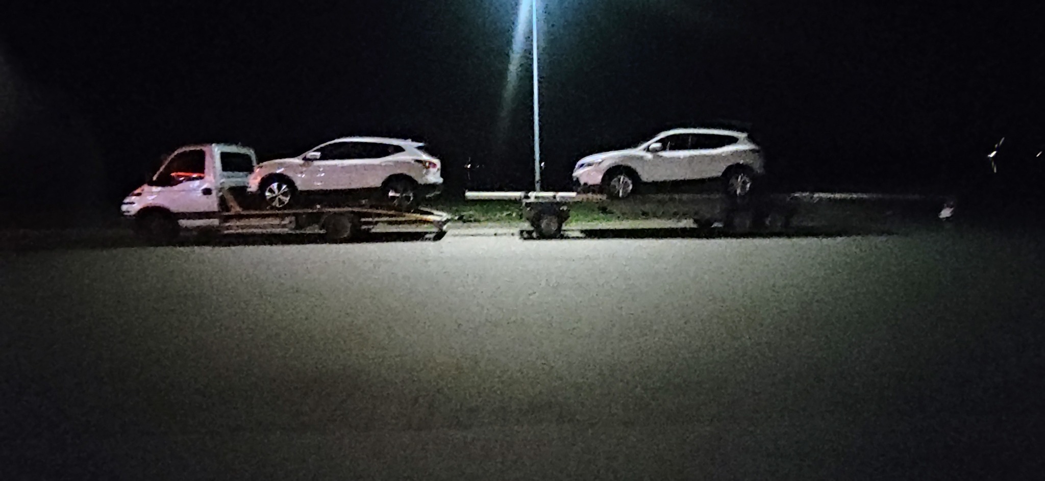 Dwa białe samochody typu SUV przewożone na lawetach w nocy, oświetlone przez uliczną latarnię.