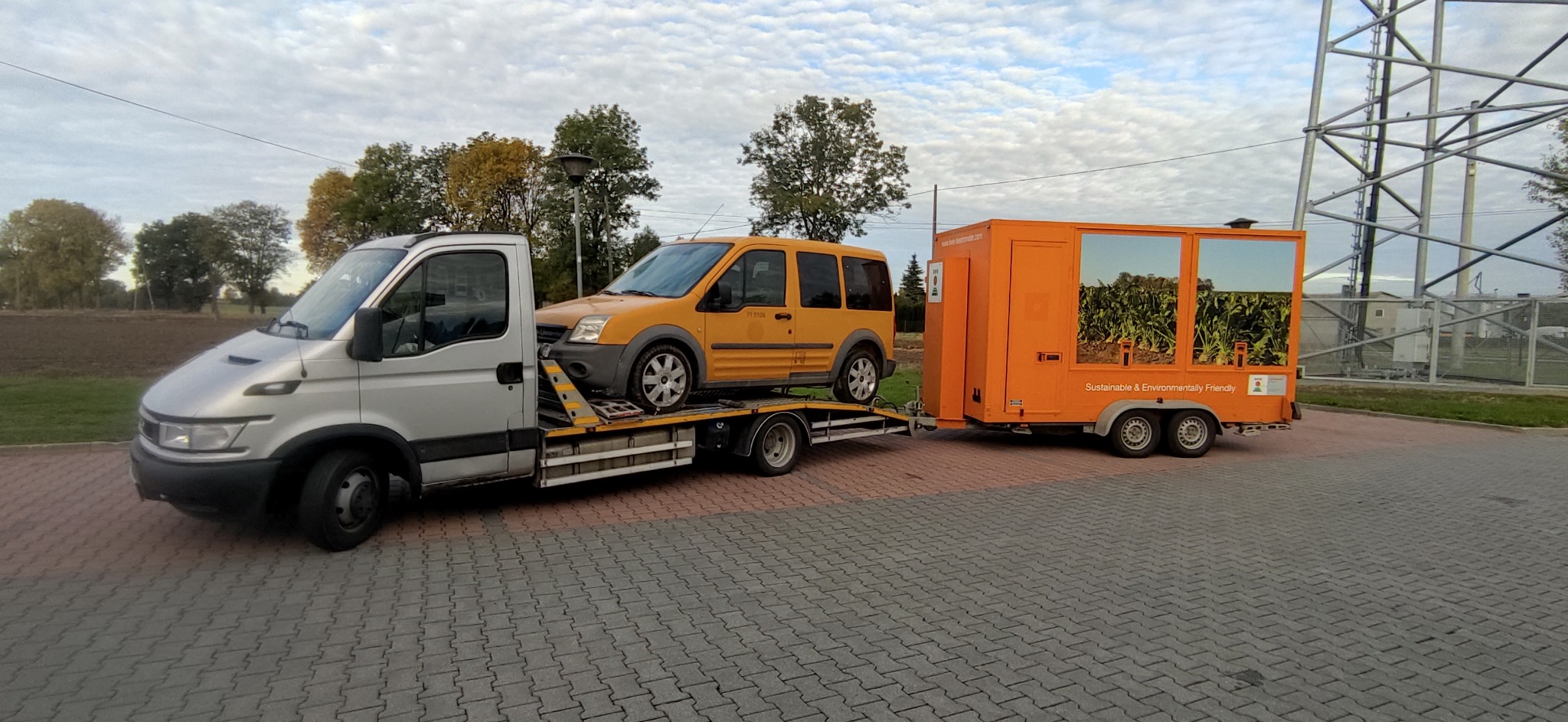 Srebrna laweta z załadowanym żółtym Fordem Connect i pomarańczową przyczepą z grafiką roślinną w tle krajobraz z drzewami i metalową konstrukcją.