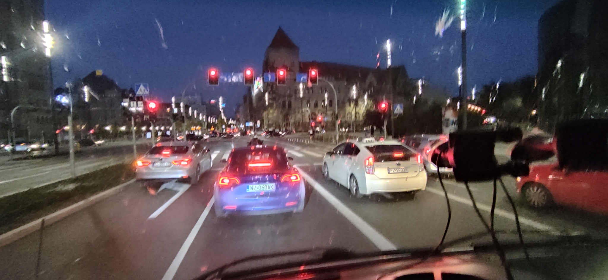 Nocny widok z wnętrza pojazdu na ulicę miejską z samochodami stojącymi na czerwonym świetle, widoczne budynki w tle i krople deszczu na szybie.