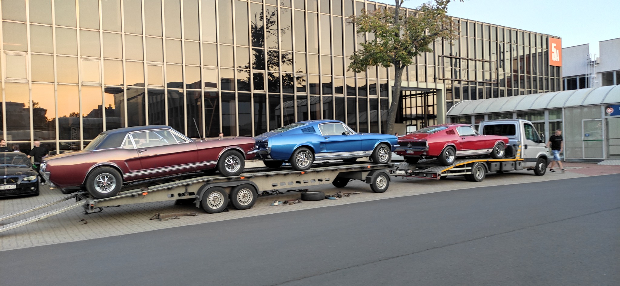 Laweta z trzema klasycznymi Ford Mustangami w różnych kolorach: bordowym, niebieskim i czerwonym, zaparkowana na tle nowoczesnego budynku z szklaną fasadą.