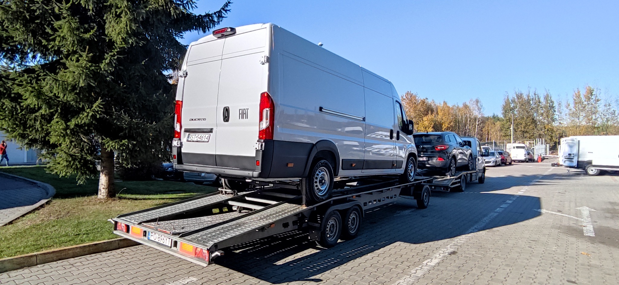 Biały Fiat Ducato na platformie lawety z czarnym samochodem osobowym za nim, w tle drzewa i inne pojazdy na parkingu