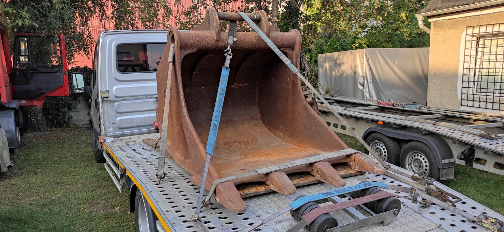 Łyżka koparki zabezpieczona pasami na platformie lawety, widoczna faktura rdzy i mocne zaczepy pasów.