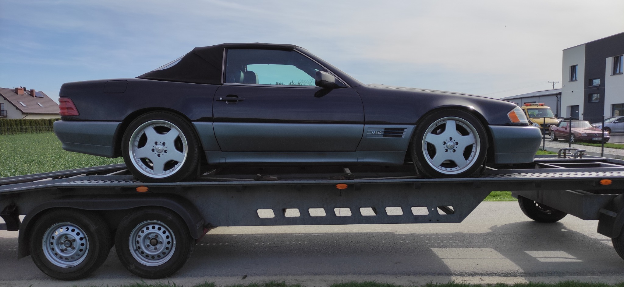 Czarny kabriolet Mercedes-Benz SL na szarej lawecie w Słupcy, widok z boku, w tle budynki i inne pojazdy.