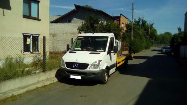 Biała laweta Mercedes-Benz Sprinter zaparkowana na ulicy w pobliżu budynku mieszkalnego i ogrodzenia z siatki.
