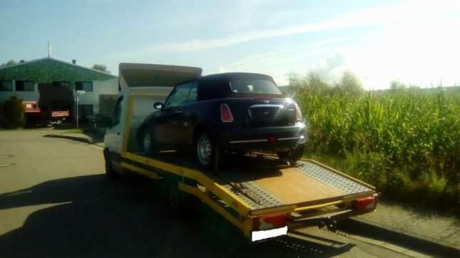 Czarny Mini Cooper Cabriolet na żółtej platformie lawety, widok z tyłu na tle budynku i pola kukurydzy.