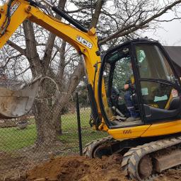 FHU Rzeszótko Tomasz - Żółta koparka JCB z operatorem w kabinie, wykonująca prace ziemne przy ogrodzeniu z siatki i drzewach bez liści w tle.