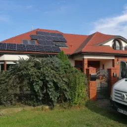 Dom jednorodzinny z czerwoną dachówką i panelami fotowoltaicznymi na dachu, częściowo zasłonięty przez zieleń, z zaparkowanym białym samochodem dostawczym z otwartymi drzwiami.