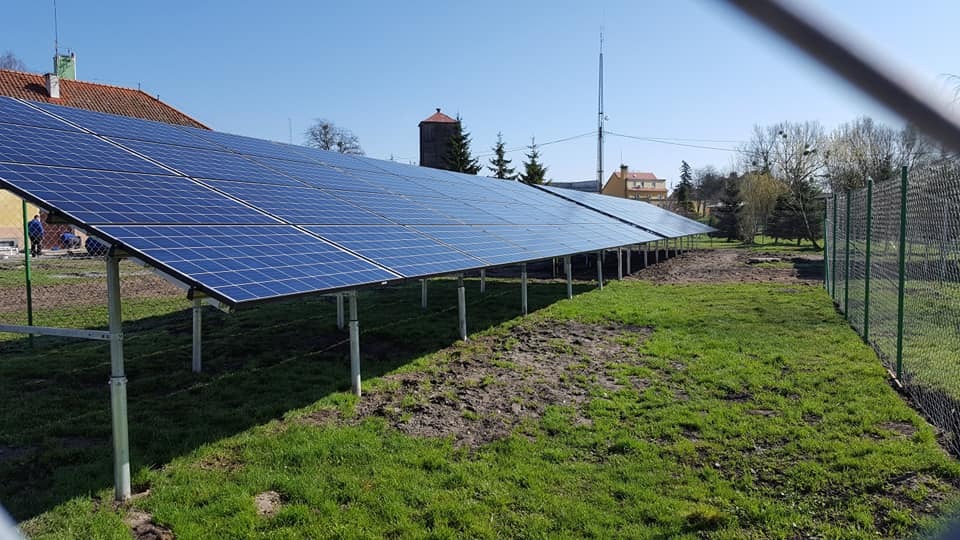 Rząd paneli słonecznych na metalowych podporach, zainstalowany na trawiastym terenie obok budynku z czerwonym dachem i wieżą ciśnień w tle, widziany zza ogrodzenia.