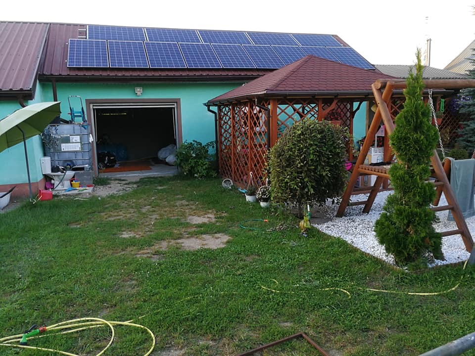 Niebiesko-zielony budynek z panelami słonecznymi na dachu, widok na podwórko z altaną, huśtawką i otwartym garażem.