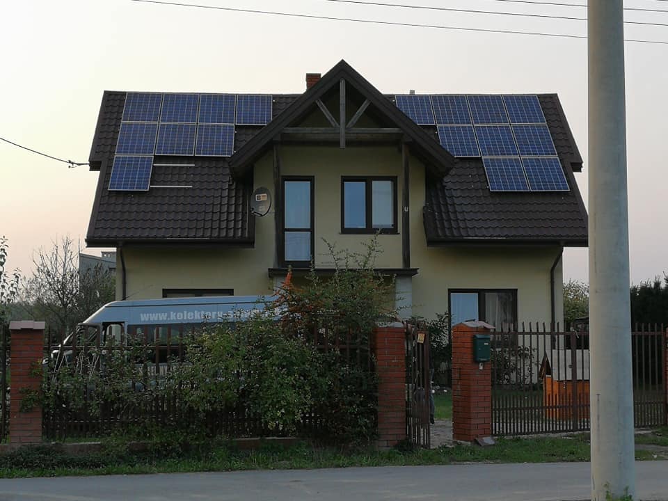 Dom jednorodzinny z zamontowanymi panelami słonecznymi na dachu, częściowo zasłonięty drzewami, z widocznym na podjeździe busem z napisem 'www.kolektory-sloneczne...'