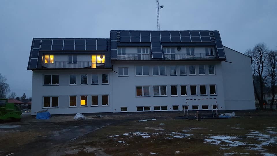 Budynek dwukondygnacyjny z panelami fotowoltaicznymi na dachu, widoczne oświetlone okna i plac budowy przed budynkiem.