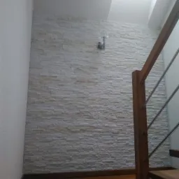 Ściana wykończona dekoracyjnym kamieniem elewacyjnym, oświetlona kinkietem, widok od dołu schodów z drewnianymi stopniami i metalową balustradą.