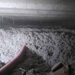 Poddasze z widoczną warstwą izolacji z włókien celulozowych, z fragmentem czerwonej rury w lewym dolnym rogu, oraz elementami konstrukcyjnymi stropu w górnej części kadru, oświetlone punktowo.