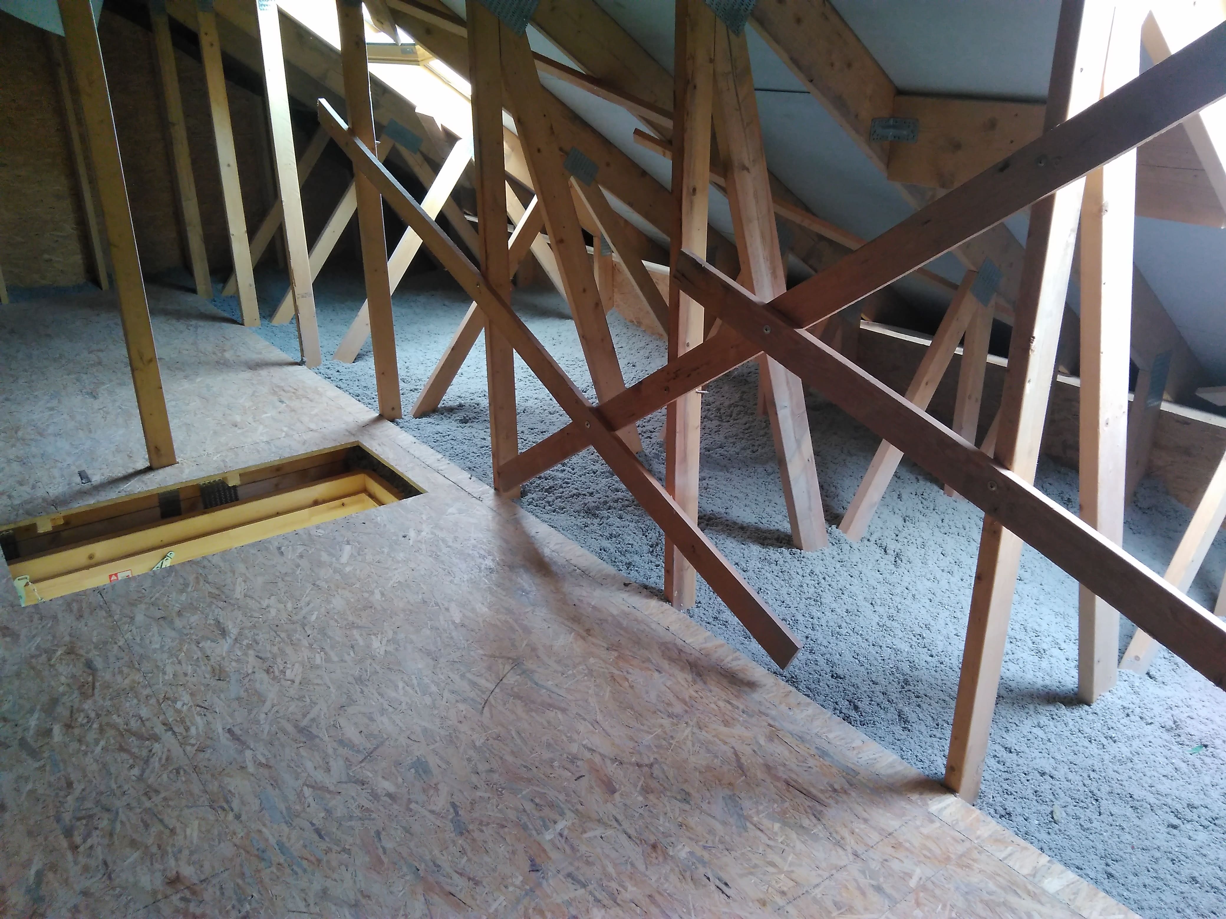 Poddasze z drewnianą konstrukcją dachu wypełnione szarą izolacją celulozową, widoczna płyta OSB na podłodze i otwór włazowy.