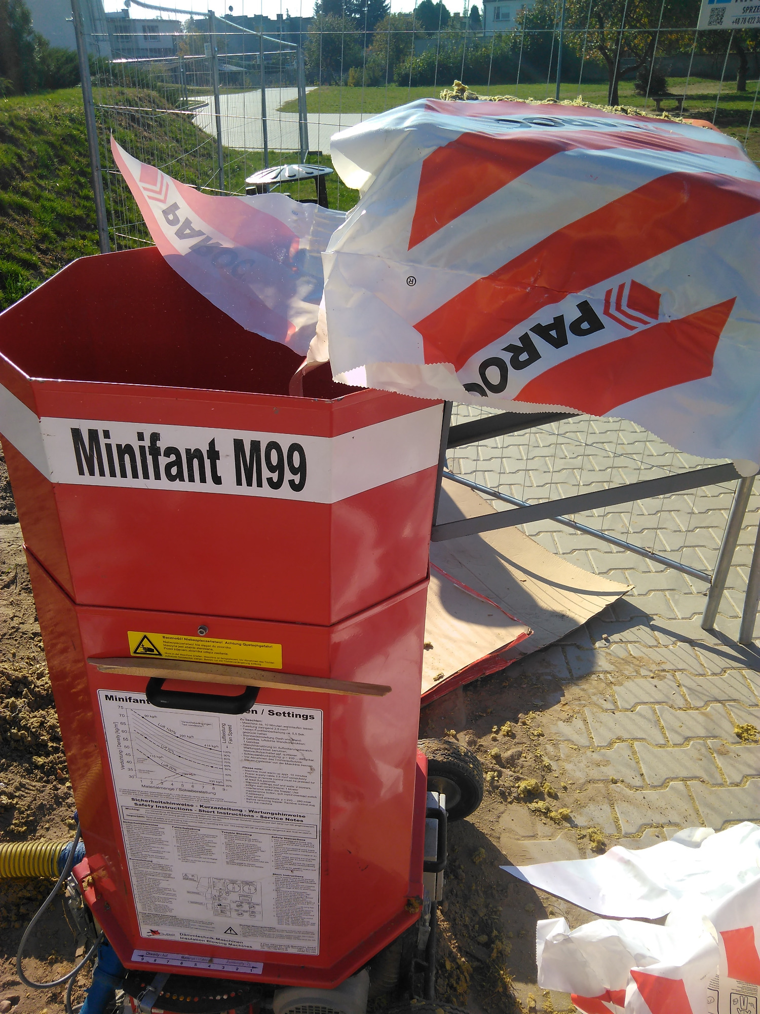 Czerwona maszyna Minifant M99 do wdmuchiwania izolacji, z widocznymi instrukcjami obsługi i workami z materiałem izolacyjnym w tle na placu budowy.