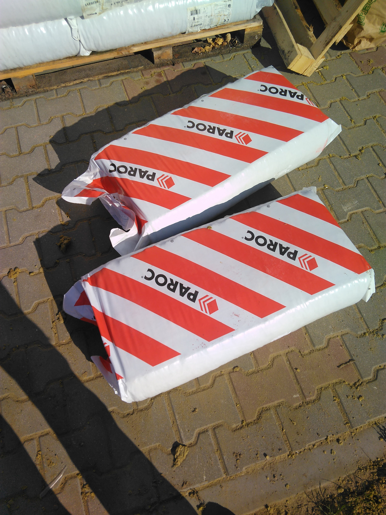 Dwa opakowania wełny mineralnej Paroc w biało-czerwone pasy leżą na brukowanej powierzchni, w tle paleta z kolejnymi opakowaniami.