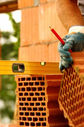 Murarz z poziomnicą i czerwoną kredką układa bloczki ceramiczne. Widoczne rękawice robocze i fragment ciała. Budowa domu w toku.