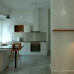 Nowoczesna, minimalistyczna kuchnia z białymi szafkami, szarym blatem i marmurowym backsplashiem. Na pierwszym planie drewniany stół z czarną nogą i designerskimi krzesłami. Ściany białe...