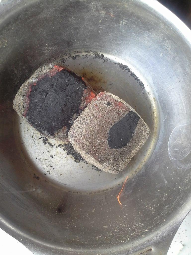 Dwa kawałki brykietu w metalowej misce, jeden zwęglony, drugi z żarzącym się brzegiem i widoczną iskrą.