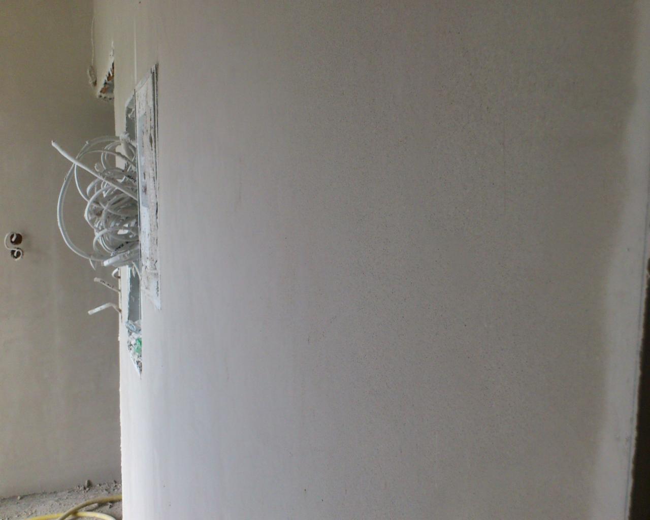 Świeżo nałożony tynk maszynowy na ścianie z widoczną puszką elektryczną i przewodami w trakcie prac wykończeniowych.