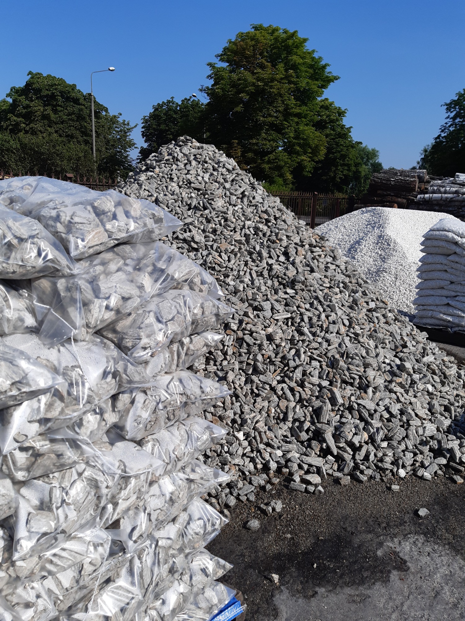 Stos szarej kory kamiennej luzem i w przezroczystych workach foliowych, na placu składowym pod błękitnym niebem.
