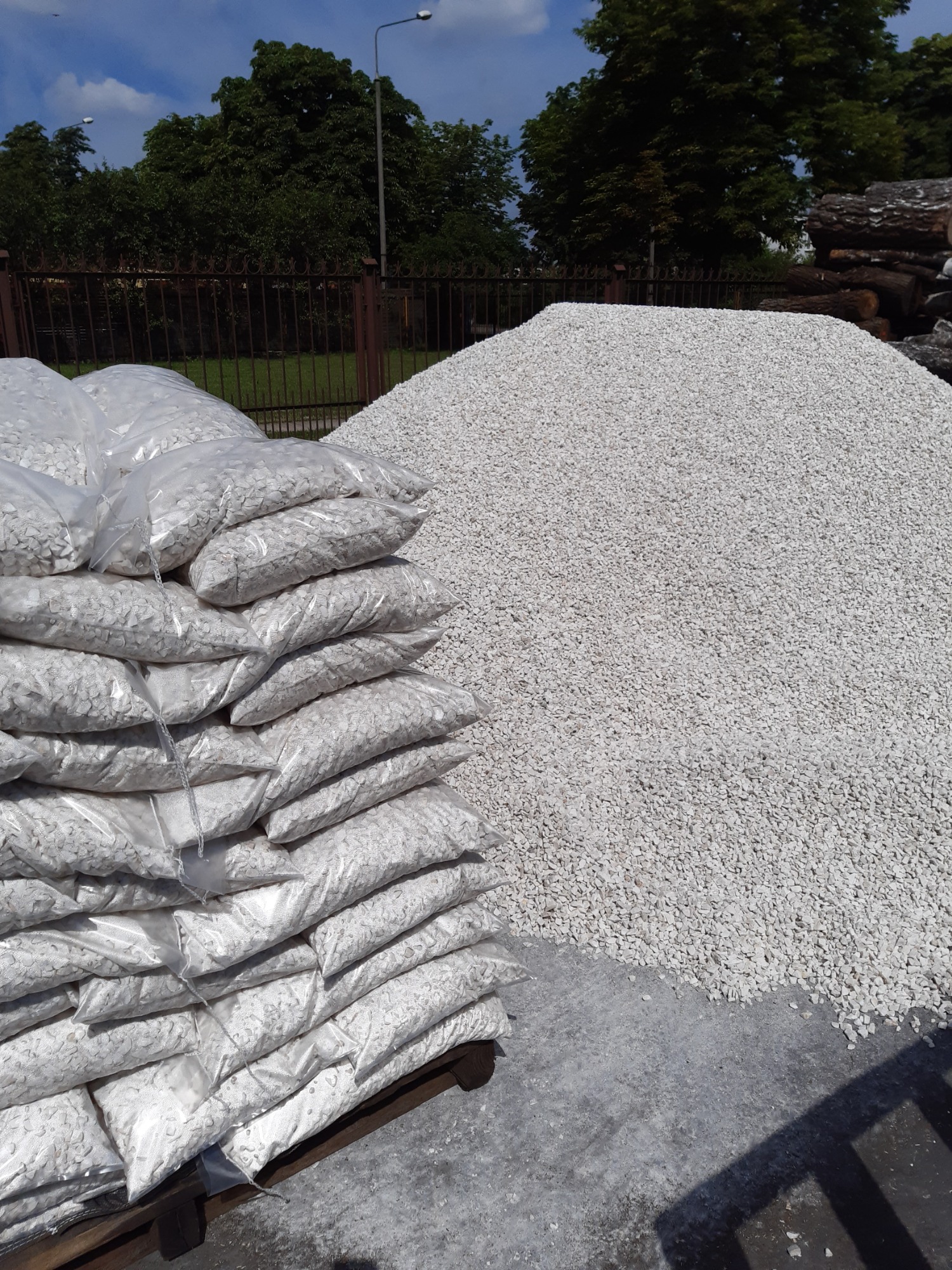 Stos worków z białym grysem na palecie obok pryzmy grysu luzem, widok na zewnątrz, w tle drzewa i ogrodzenie.