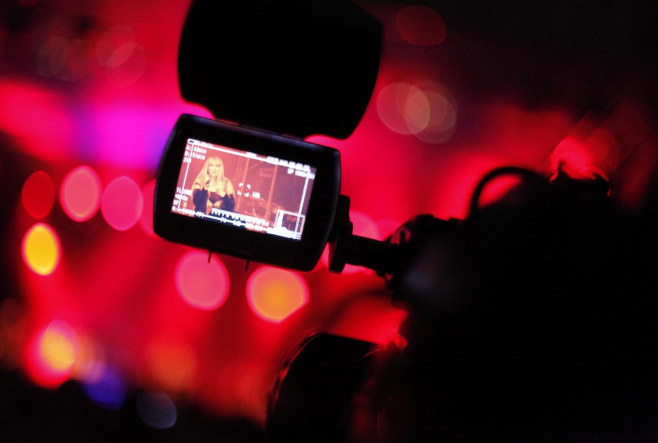 Ekran kamery filmowej z widoczną blond prezenterką na tle rozmytych czerwonych świateł scenicznych.