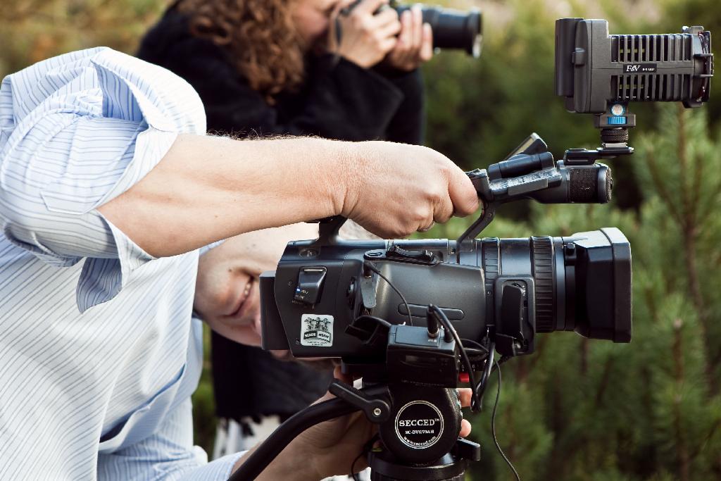 Operatorzy kamer w trakcie pracy, jeden z profesjonalną kamerą wideo na statywie Secced, drugi z aparatem fotograficznym z teleobiektywem, na tle zieleni.