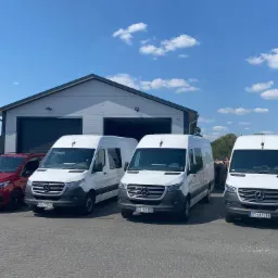 Flota pojazdów, składająca się z trzech białych vanów i jednego czerwonego samochodu osobowego marki Mercedes-Benz, zaparkowana przed szarym budynkiem z bramami garażowymi w słoneczny dzień.