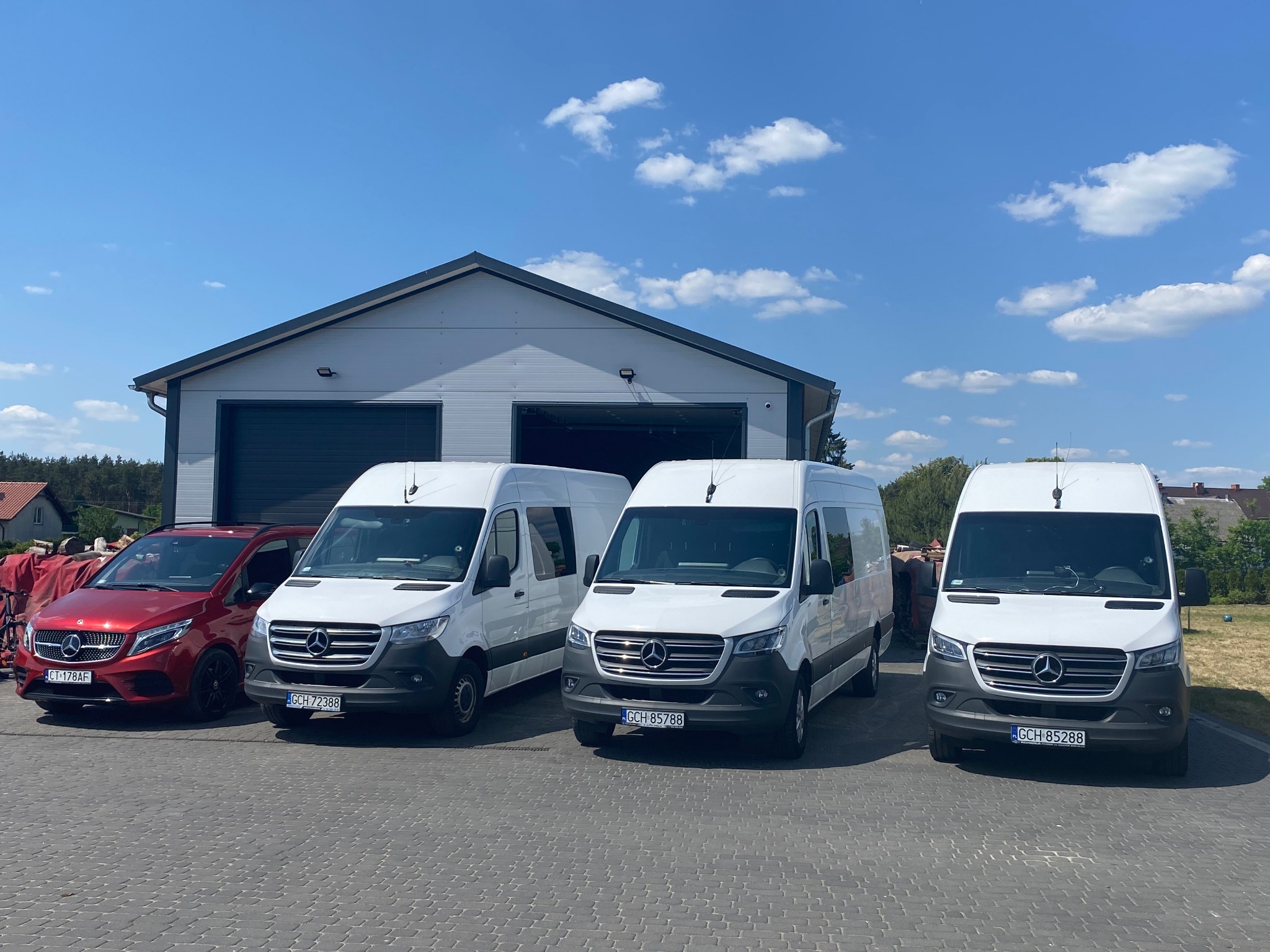 Flota pojazdów, składająca się z trzech białych vanów i jednego czerwonego samochodu osobowego marki Mercedes-Benz, zaparkowana przed szarym budynkiem z bramami garażowymi w słoneczny dzień.