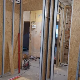 Remont mieszkania w trakcie: widoczne stelaże ścian z płyt OSB, narzędzia (wiertarka) i materiały izolacyjne. Przewody elektryczne, surowy stan ścian w tle.